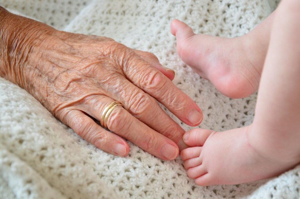 Main d’une personne âgée posée près des pieds d’un bébé, évoquant la transmission entre générations