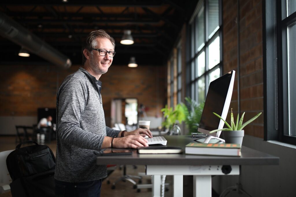 Homme senior aux cheveux gris, souriant, travaillant debout devant un ordinateur dans un bureau lumineux